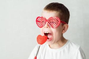 retrato de un niño con gafas en forma de corazón en una camiseta blanca contra un fondo blanco mordiendo un corazón. contra el dia de san valentin foto