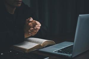 concepto de tecnología cristiana en línea. manos rezando de cristiano con computadora portátil digital, iglesia en vivo en línea para el servicio dominical. el hombre católico asiático está leyendo el libro sagrado de la biblia y estudiando en línea. foto