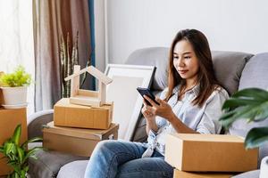 Happy young asian woman using smartphone in living room at new house with stack of cardboard boxes on moving day photo