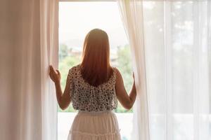 young woman opening curtains in the morning and looking through the window photo