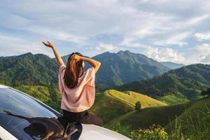 Young woman traveler sitting on a car watching a beautiful mountain view while travel driving road trip on vacation