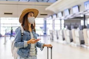 Young woman wearing a face mask and holding a mobile phone is looking for a check-in counter at the airport, New normal lifestyle concept photo