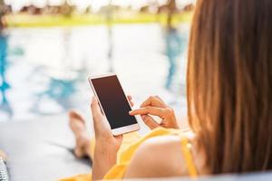 Young woman traveler relaxing and using a mobile phone by a hotel pool while traveling for summer vacation photo