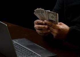 Computer and hands of a man wearing a black shirt, sitting on a chair and a table, is a thief, holding money, counting the amount obtained from hijacking or robbing, in a pitch-black room. photo