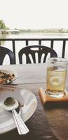 cuchara en un plato blanco con comida y mezcla de whisky tailandés frío con soda y agua en una mesa de madera con fondo de vista al lago o al río en estilo de color vintage. relajarse y beber a la hora del almuerzo concepto foto