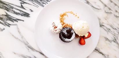 Flat lay or Top view of Chocolate Lava Cake with vanilla ice cream, whipped cream and sliced of strawberry in plate on white marble table. Dessert and Sweet food concept photo