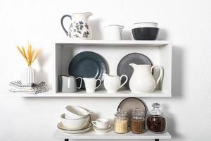 vista frontal de la cocina hermosos platos de cerámica y porcelana en estantes blancos. tarros de cristal con productos sueltos, especias. pared blanca. foto
