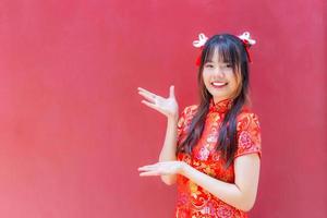 bella y bonita jovencita asiática con el pelo largo que lleva un vestido rojo de cheongsam con el tema del año nuevo chino mientras muestra su mano para presentar algo de fondo rojo. foto