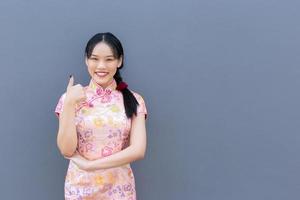 Asian woman with long hair who wears pink Cheongsam dress in Chinese new year theme while her hand shows to present something and showing hand as thump up smiling happily with grey background. photo