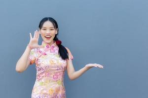 hermosa mujer asiática con el pelo largo que usa un vestido rosa cheongsam en el tema del año nuevo chino mientras su mano muestra algo y muestra la mano para señalar un fondo gris foto