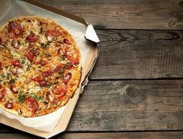 baked round pizza with smoked sausages, mushrooms, tomatoes, cheese and dill in an open cardboard box on a wooden table photo