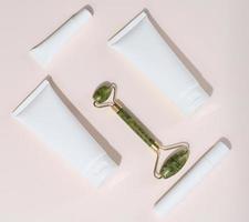 Jade manual facial massager and white plastic tubes for cosmetics on a beige background photo