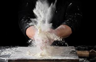 chef in black uniform sprinkles white wheat flour over the table photo