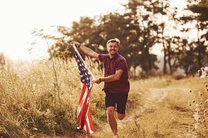 Adult man runs with American Flag in hands outdoors in the field. Feels freedom at sunny daytime photo