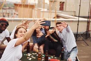 Girl doing photo when people playing card game. Group of young people in casual clothes have a party at rooftop together at daytime
