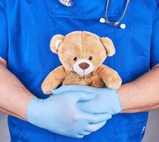 médico con uniforme azul y guantes de látex sosteniendo un oso de peluche marrón foto