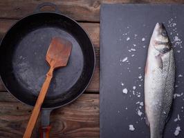 fresh whole sea bass fish on a black board, next to it is an empty round black frying pan photo