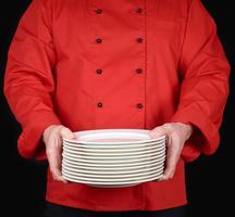 cook in red uniform holds in his hands a stack of round white empty plates, black photo
