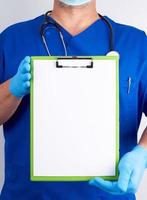 médico con uniforme azul y guantes de látex sostiene un soporte verde para hojas de papel foto