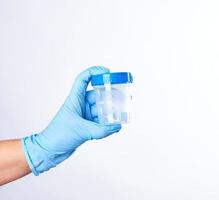 la mano en un guante estéril azul sostiene un recipiente de plástico para recolectar análisis foto