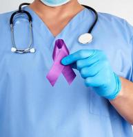 doctor in blue uniform and latex gloves holds a purple ribbon as a symbol of early research and disease control photo