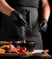 chef in black uniform and latex gloves sprinkles with white salt raw chicken meat in a black cast-iron frying pan photo