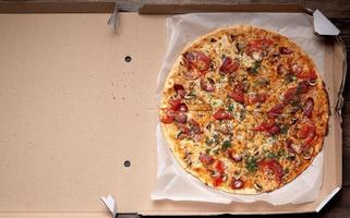 baked round pizza with smoked sausages, mushrooms, tomatoes, cheese and dill in an open cardboard box photo