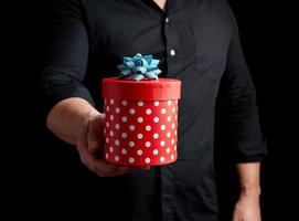 adult man in a black shirt holds in his hand a round red box of polka dots with a blue bow photo