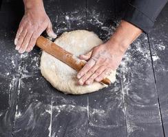 chef in a black tunic rolls a dough for a round pizza photo