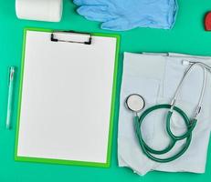 paper holder with empty white sheets, medical stethoscope, pills on a green background photo