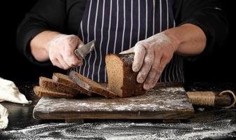 chef con uniforme negro sostiene un cuchillo de cocina en la mano y corta trozos de pan de una hogaza de harina de centeno marrón horneada foto