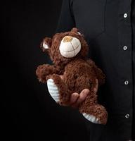 adult man in a black shirt holds a brown teddy bear on a dark background photo