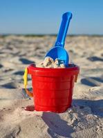 Baby red plastic bucket with sand and shovel on the seashore photo