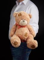 man in a white shirt holds a large teddy beige bear with patches photo