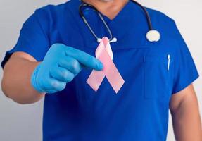 doctor in blue uniform and sterile latex gloves holds a pink ribbon a symbol of the fight against breast cancer photo