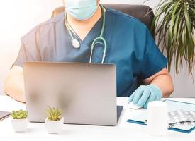 médico masculino sentado en una mesa blanca con uniforme azul y guantes de látex, el especialista trabaja en una laptop foto