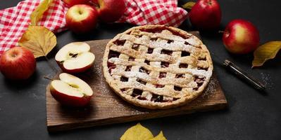 baked round traditional apple pie on brown wooden board and fresh red apples, top view photo