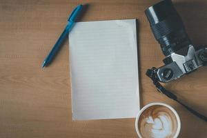 vista superior de papel vacío, taza de café, cámara y bolígrafo en la mesa de madera foto