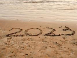 foto de concepto de año nuevo. números 2023 escritos a mano en la superficie de la arena. ola de mar suave y playa en el fondo.