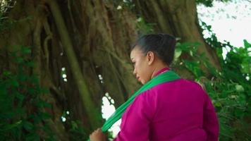 An Asian woman wearing a green scarf in a traditional pink dress after doing a ritual in front of the big tree inside the forest