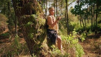Balinese man resting and leaning on the tree while wearing a golden crown and shirtless inside the jungle video
