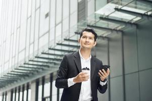 Handsome smiling asian businessman holding smartphone in office Businessman looking at hand holding coffee cup while standing alone in modern workplace. photo