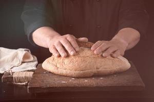 las manos masculinas sostienen pan de centeno horneado marrón sobre una tabla de madera foto