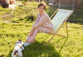 Young woman in pajama resting in chair on green lawn on sunny summer day and pet jack russell terrier dog - village and country life photo