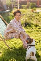 Young woman in pajama resting in chair on green lawn on sunny summer day and pet jack russell terrier dog - village and country life photo