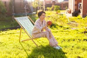 Young woman in pajama is resting in chair on a green lawn on sunny summer day - village and country life photo