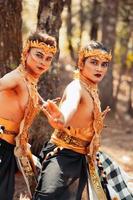 Two Balinese men dancing together in front of the tree while wearing a golden crown and golden necklaces in stripped clothes photo