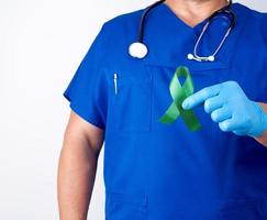 doctor in blue uniform and latex gloves holds a green ribbon as a symbol of early research and disease control photo