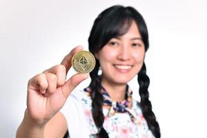 retrato de una hermosa joven asiática feliz vestida de denim sosteniendo una moneda criptográfica sobre fondo blanco. foto