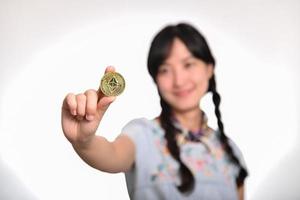 Portrait of beautiful happy young asian woman in denim dress holding crypto currency coin on white background. photo